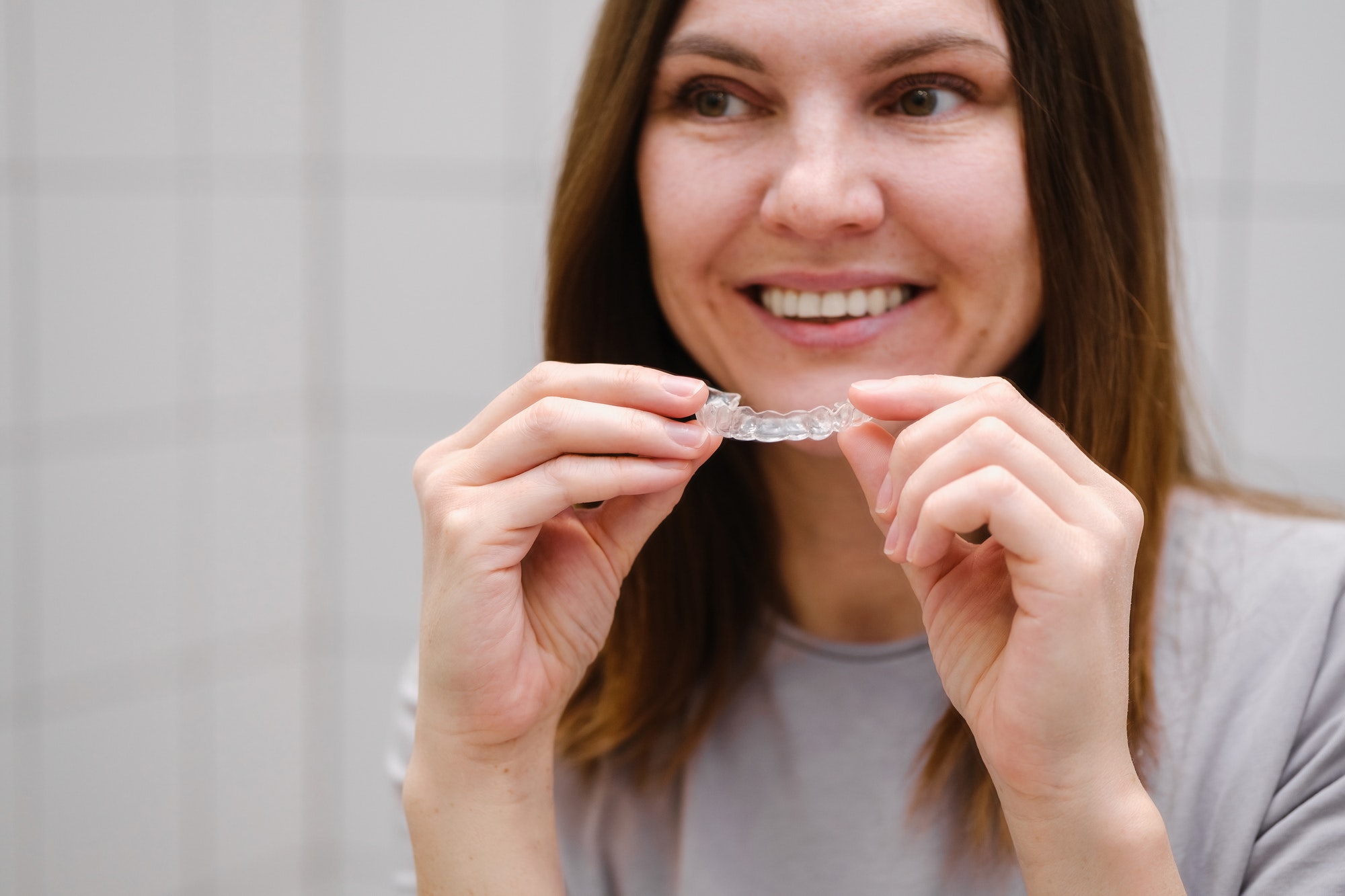 A woman in front of a mirror holding invisible plastic teeth aligners in hands. Putting on braces.